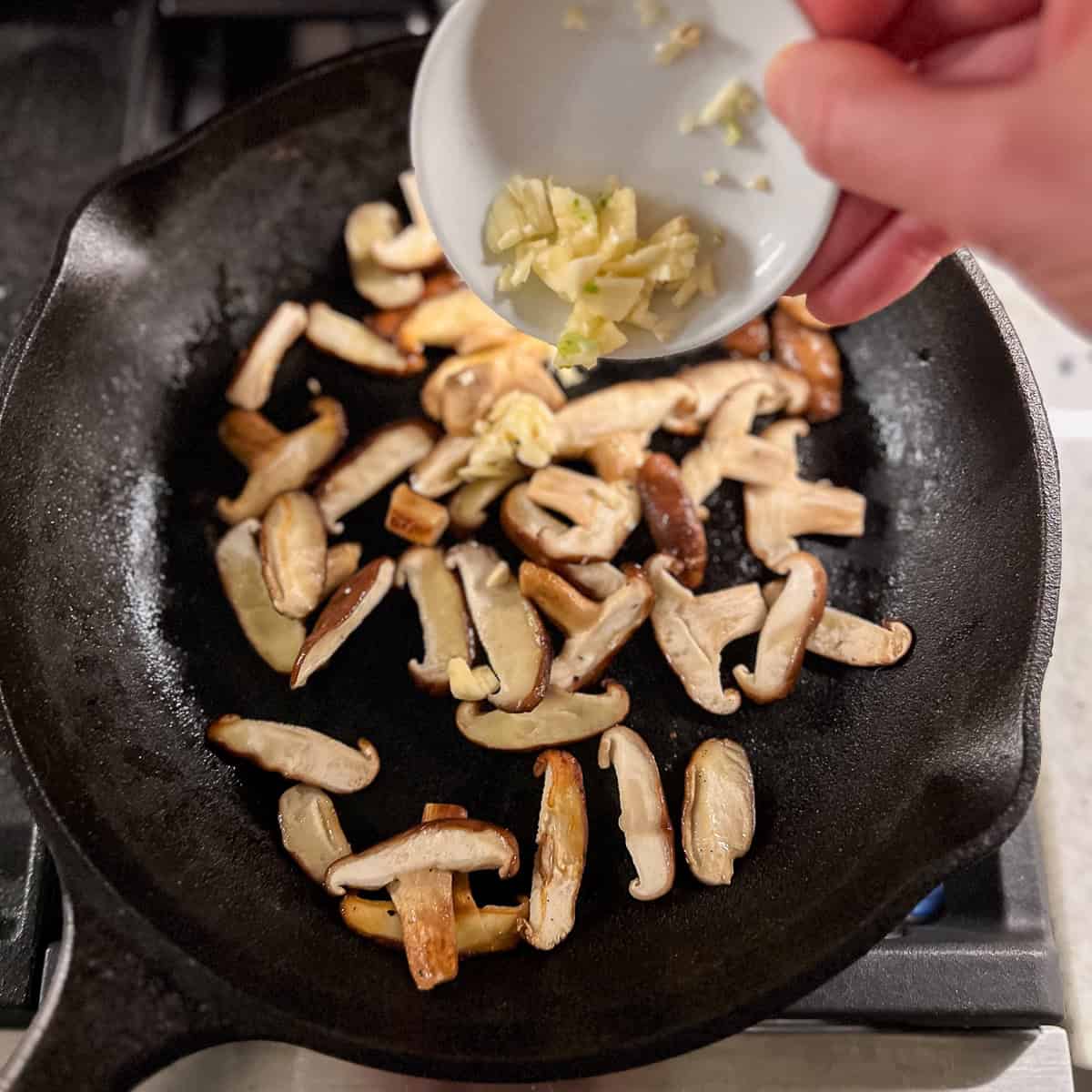 Sliced mushrooms sautéed in a skillet with minced garlic being added.