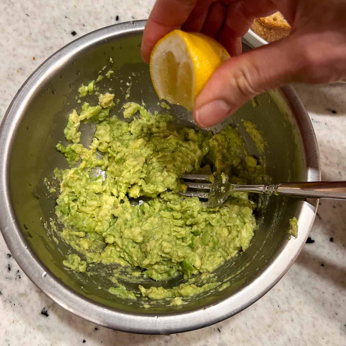 Smashed avocado in a mixing bowl with fresh squeezed lemon juice being added.