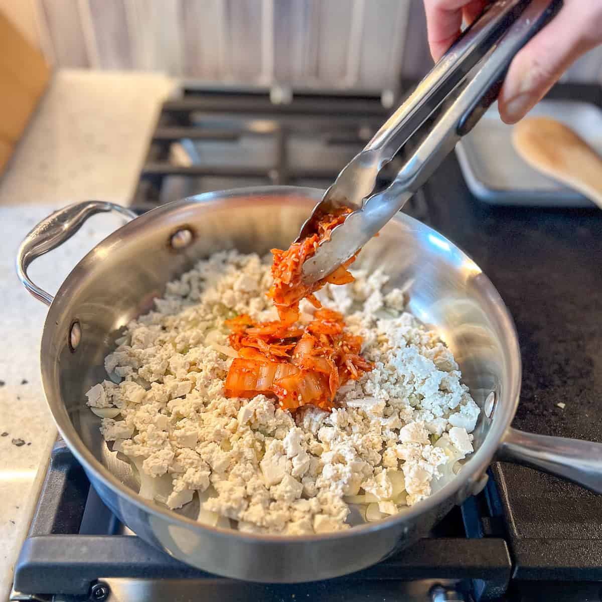 Kimchi being added to the skillet with crumbled tofu, onion and garlic.