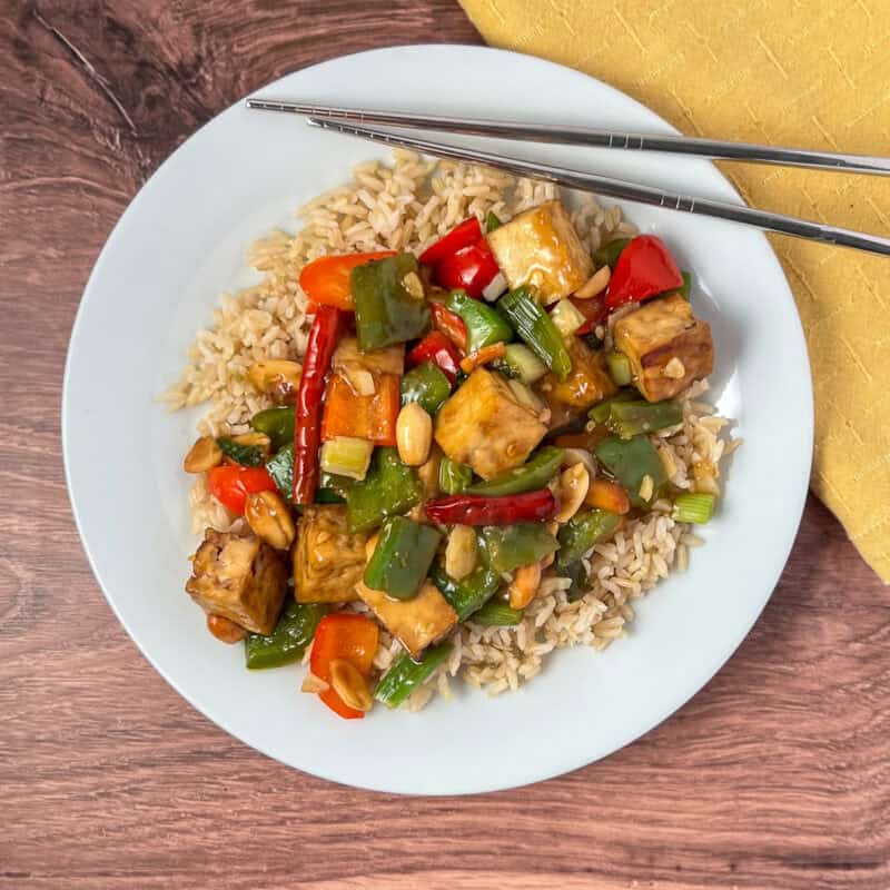 Kung pao tofu over brown rice on a plate with chopsticks.