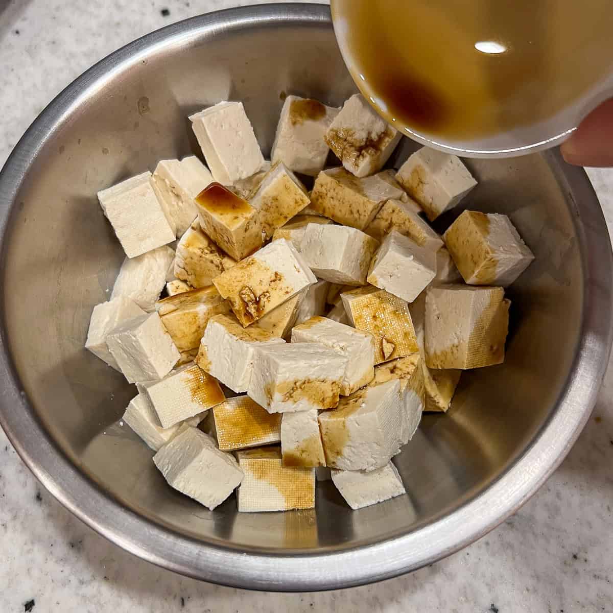 Coconut aminos being added to a bowl of tofu chunks.