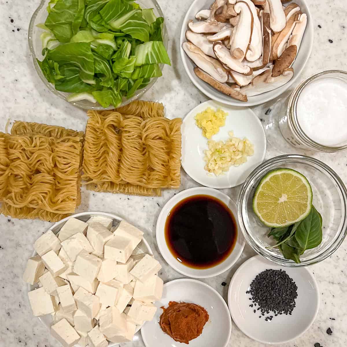 Ingredients for curry ramen: ramen noodles, tofu, mushrooms, baby bok choy, ginger, garlic, coconut milk, coconut aminos, red curry paste, Thai basil, lime, and black sesame seeds.