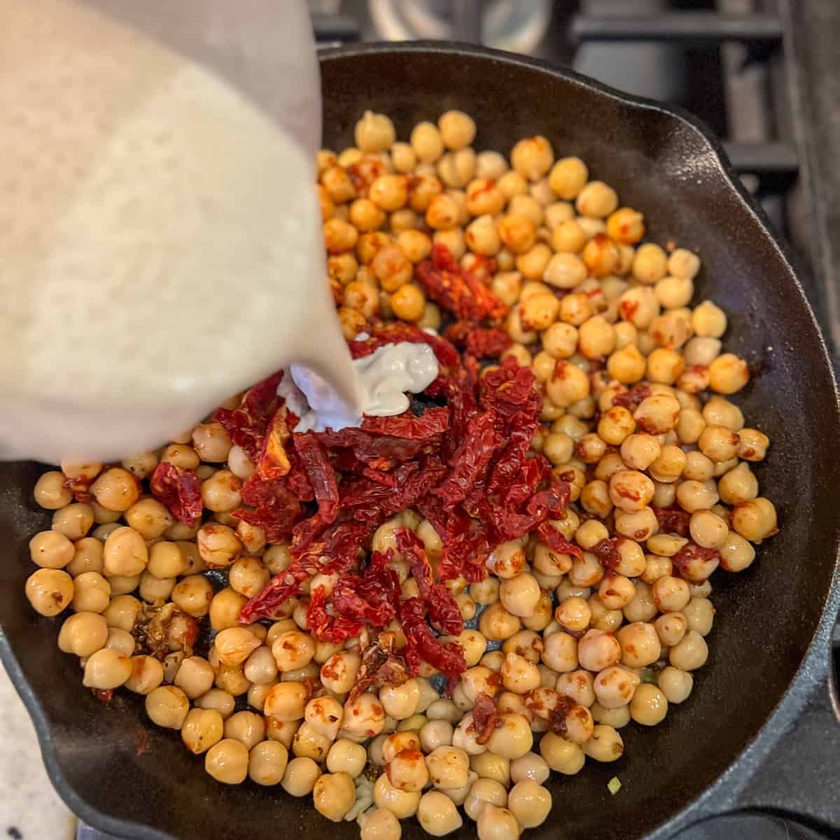 Cashew cream being poured into the skillet with chickpeas and sundried tomatoes.