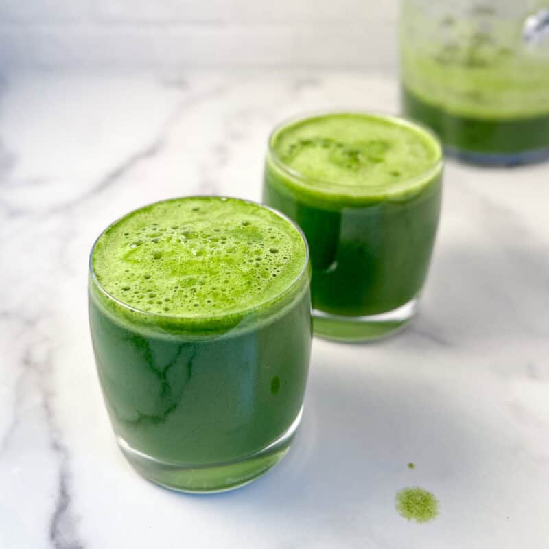 Two glasses of super green juice with pitcher blurred in the background.