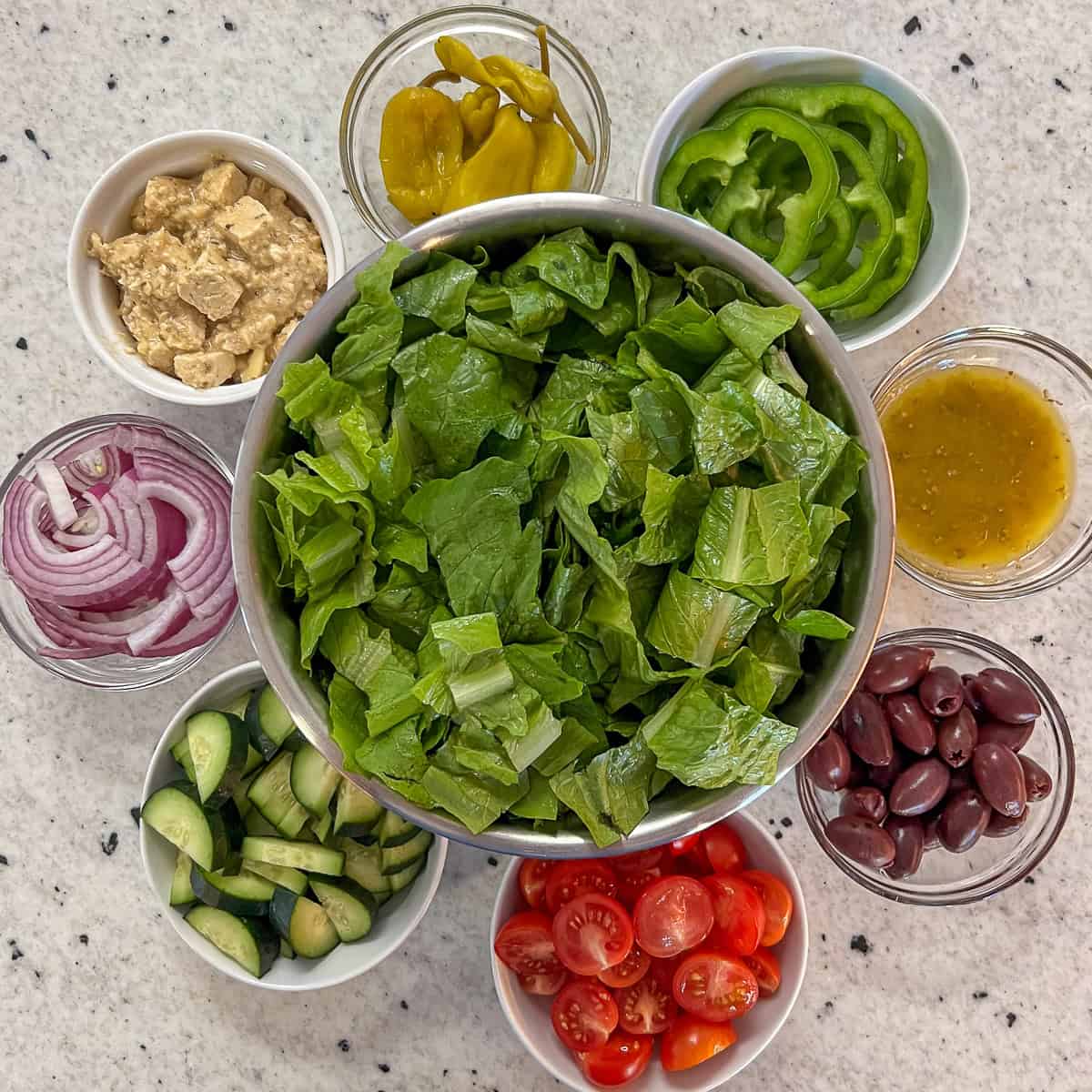 Greek salad ingredients: romaine lettuce, cherry tomatoes, pitted kalamata olives, cucumbers, red onion, green bell pepper, pepperoncini, vegan feta and Greek salad dressing.