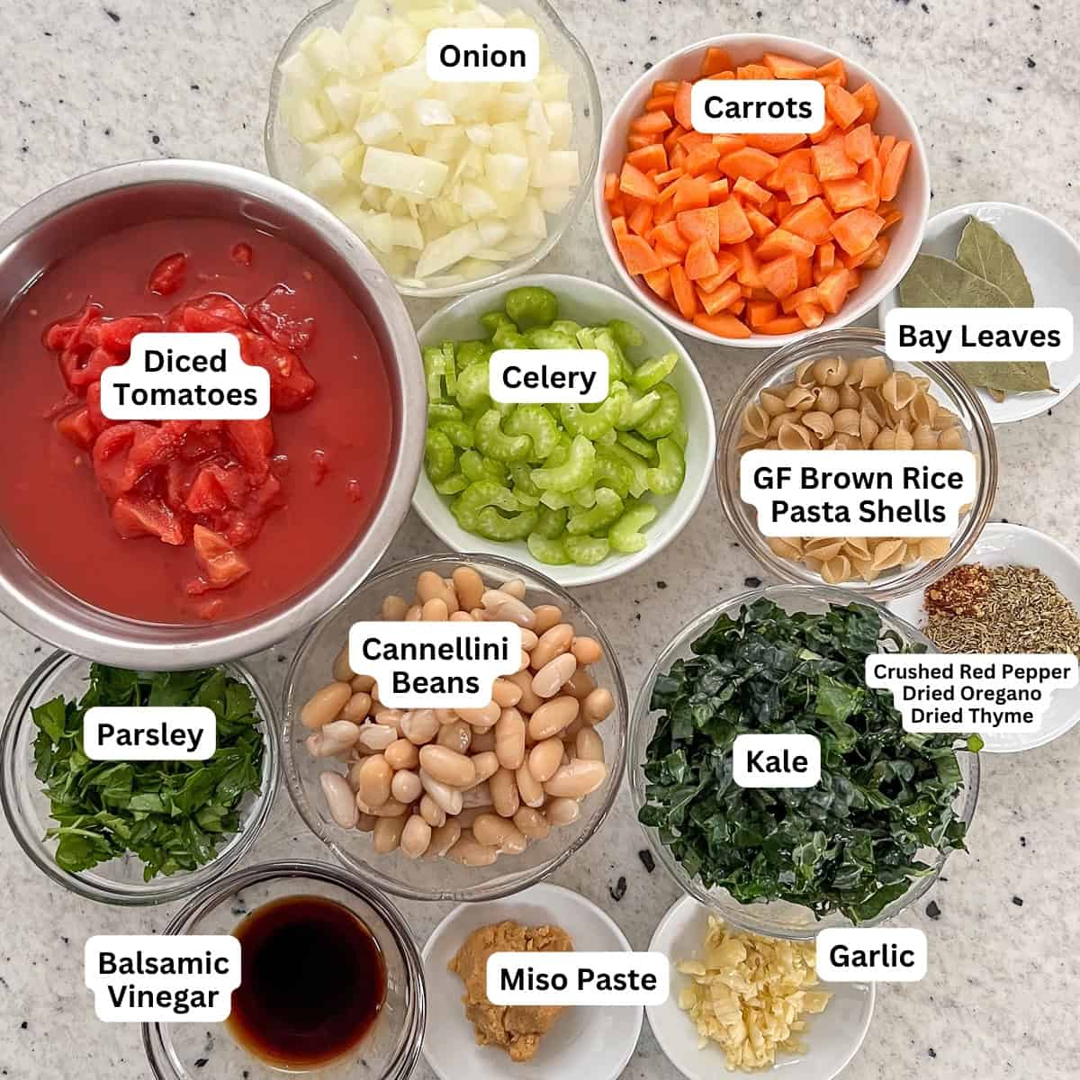 Ingredients for Vegan Minestrone soup: onion, carrots, celery, small shells pasta, bay leaves, garlic, seasonings, kale, cannellini beans, miso paste, balsamic vinegar, parsley, diced tomatoes, and chopped kale.