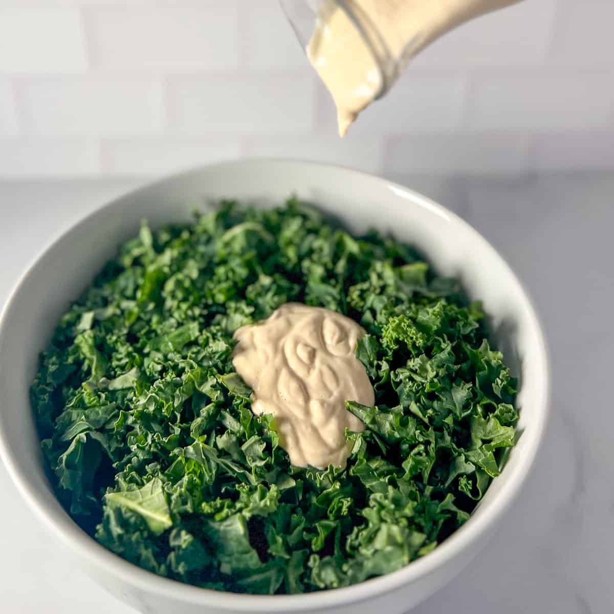 Tahini lemon garlic sauce being added to a bowl of chopped kale.