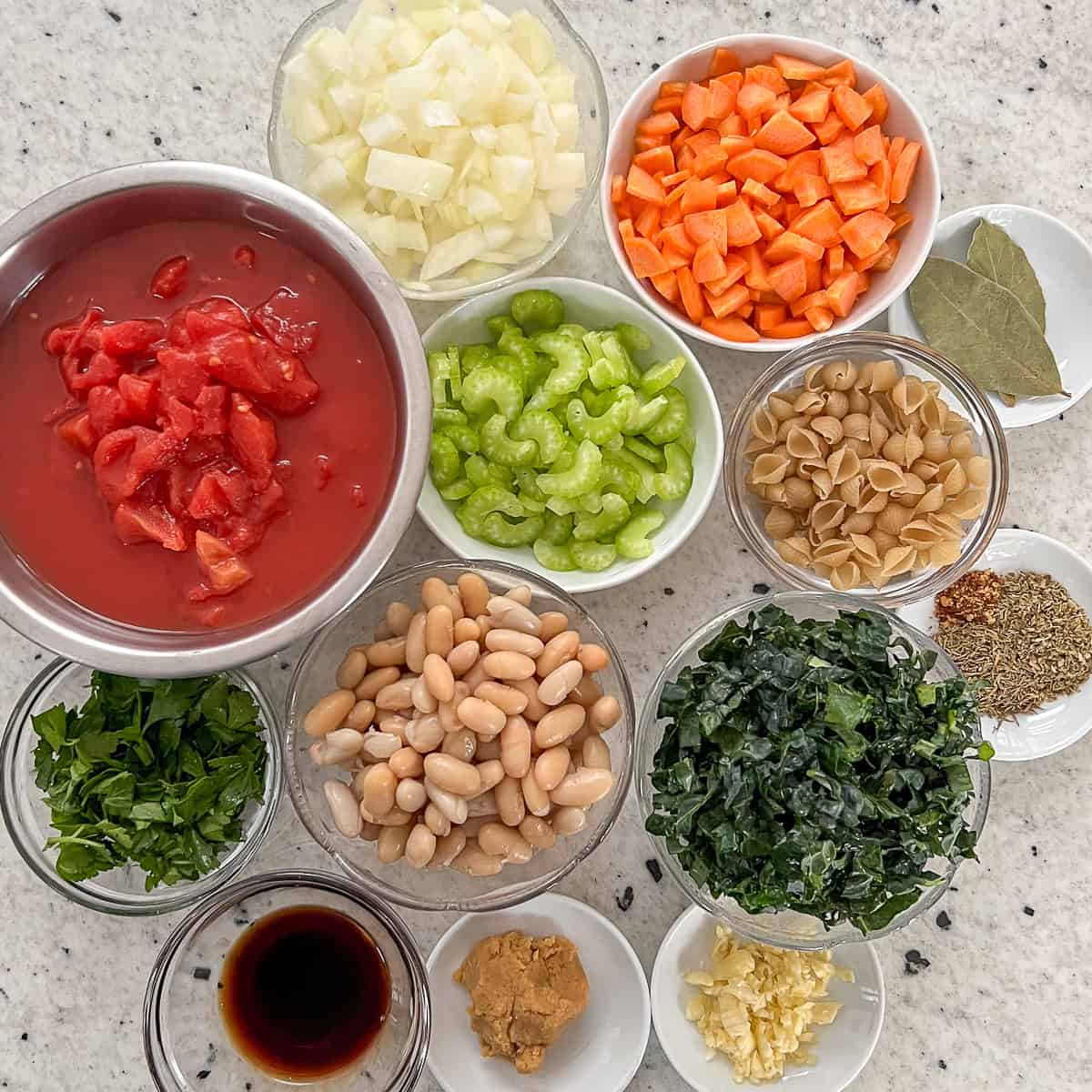 Ingredients for Vegan Minestrone soup: onion, carrots, celery, small shells pasta, bay leaves, garlic, seasonings, kale, cannellini beans, miso paste, balsamic vinegar, parsley, diced tomatoes, and chopped kale.