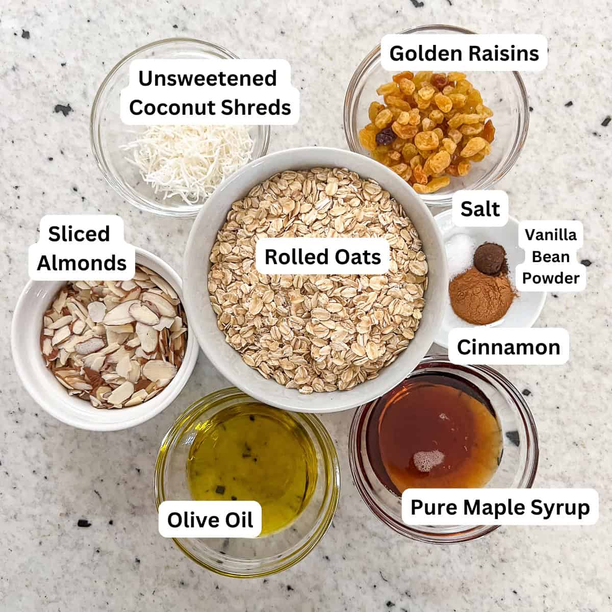 Ingredients for vanilla almond granola: rolled oats, unsweetened shredded coconut, golden raisins, sliced almonds, cinnamon, vanilla bean powder, salt, maple syrup and olive oil.