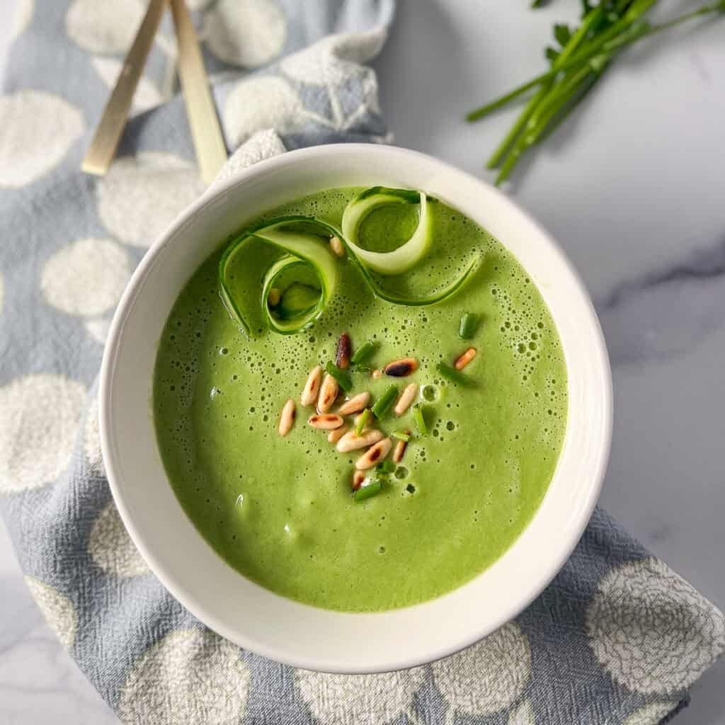 A bowl of cucumber gazpacho soup in a bowl topped with shaved cucumber and toasted pinenuts.