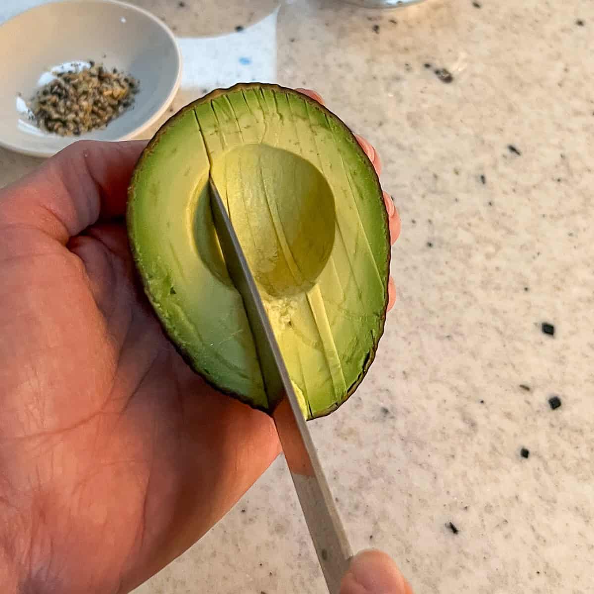 A woman holding the avocado in one hand and using a sharp knife to cut thin strips.