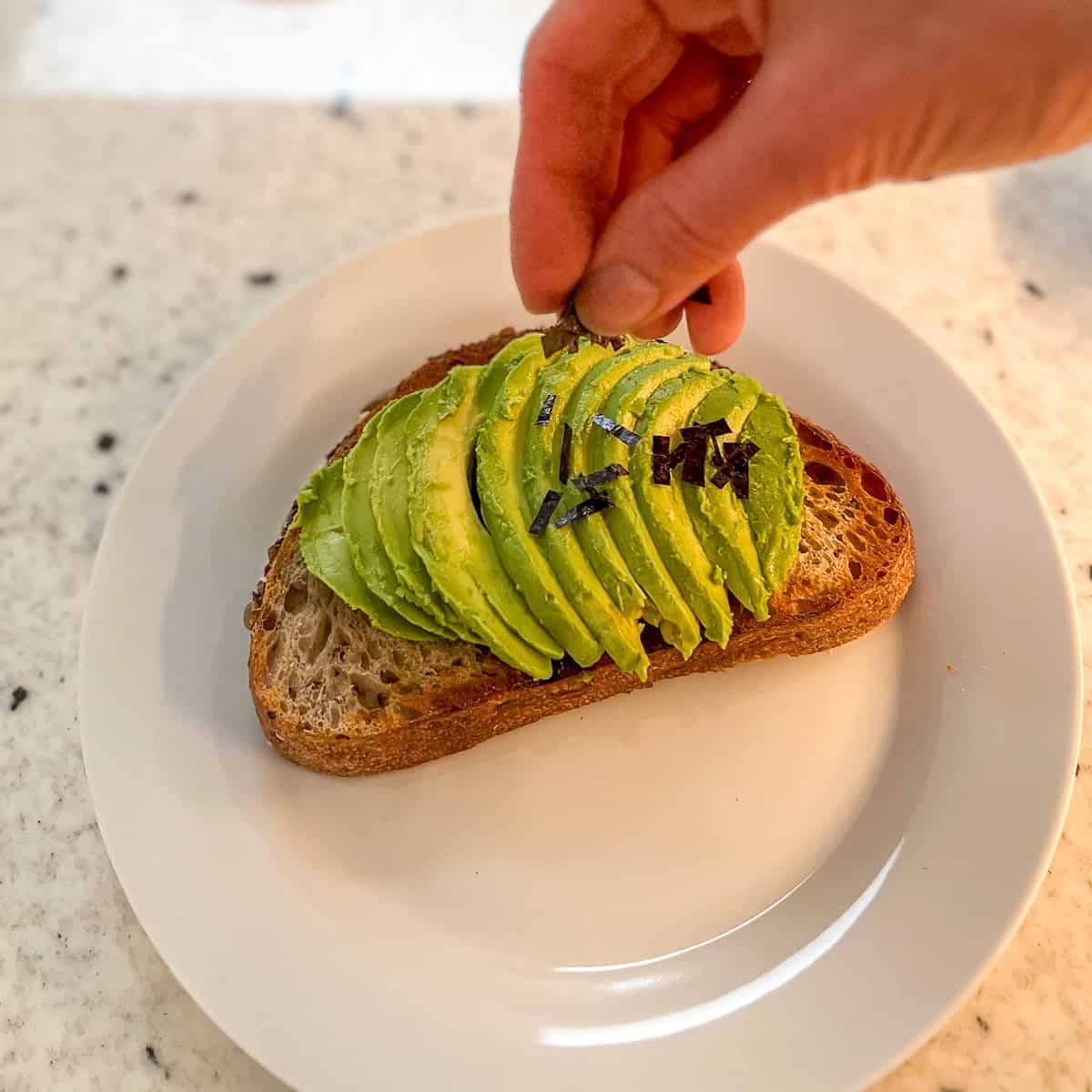 A woman's hand sprinkling minced nori seaweed across the avocado toast.