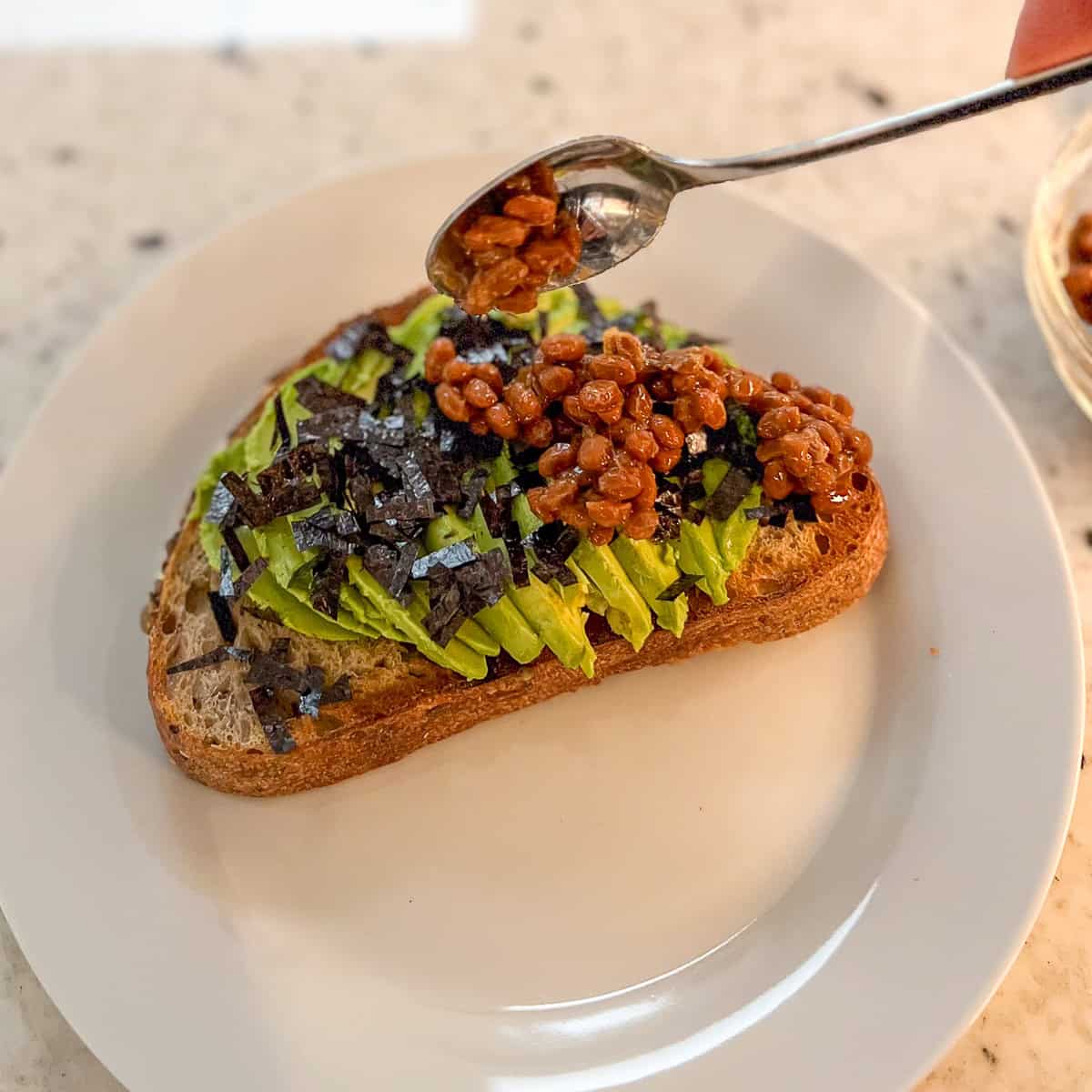 Natto being added to the avocado toast with a spoon.