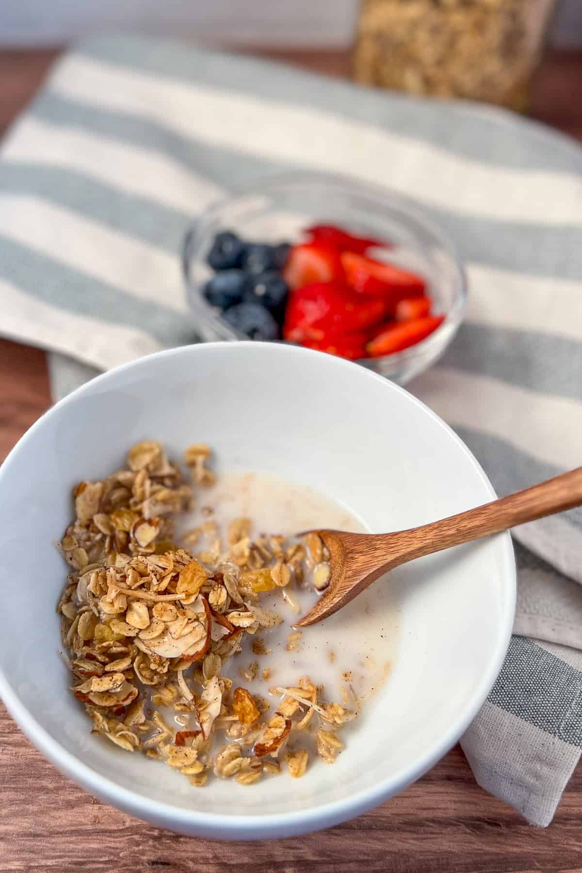 Vanilla Almond Granola in a bowl with almond milk and a wooden spoon; bowl of berries blurred in the background.