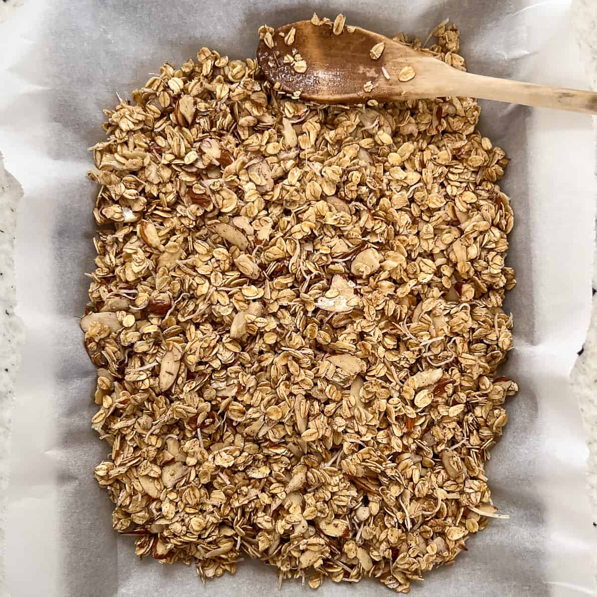 Vanilla almond granola evenly spread on baking sheet and patted down with a wooden spoon.