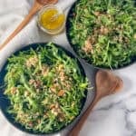 Two bowls of arugula quinoa salad with salad tongs on the side and dressing.