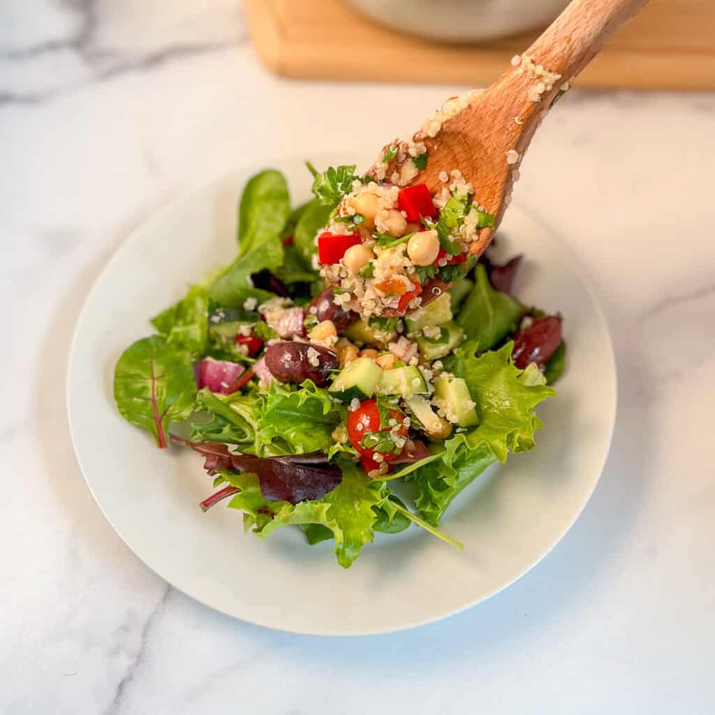 A wooden spoon adding Mediterranean quinoa salad over a bed of mixed salad greens on a plate.