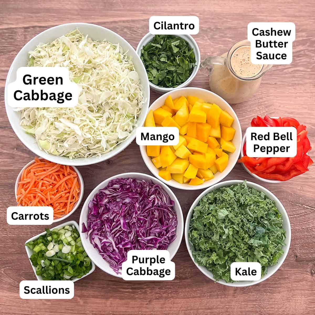 Individual bowls of green cabbage, cilantro, cashew sauce, purple cabbage, kale, carrots, mango and scallions.