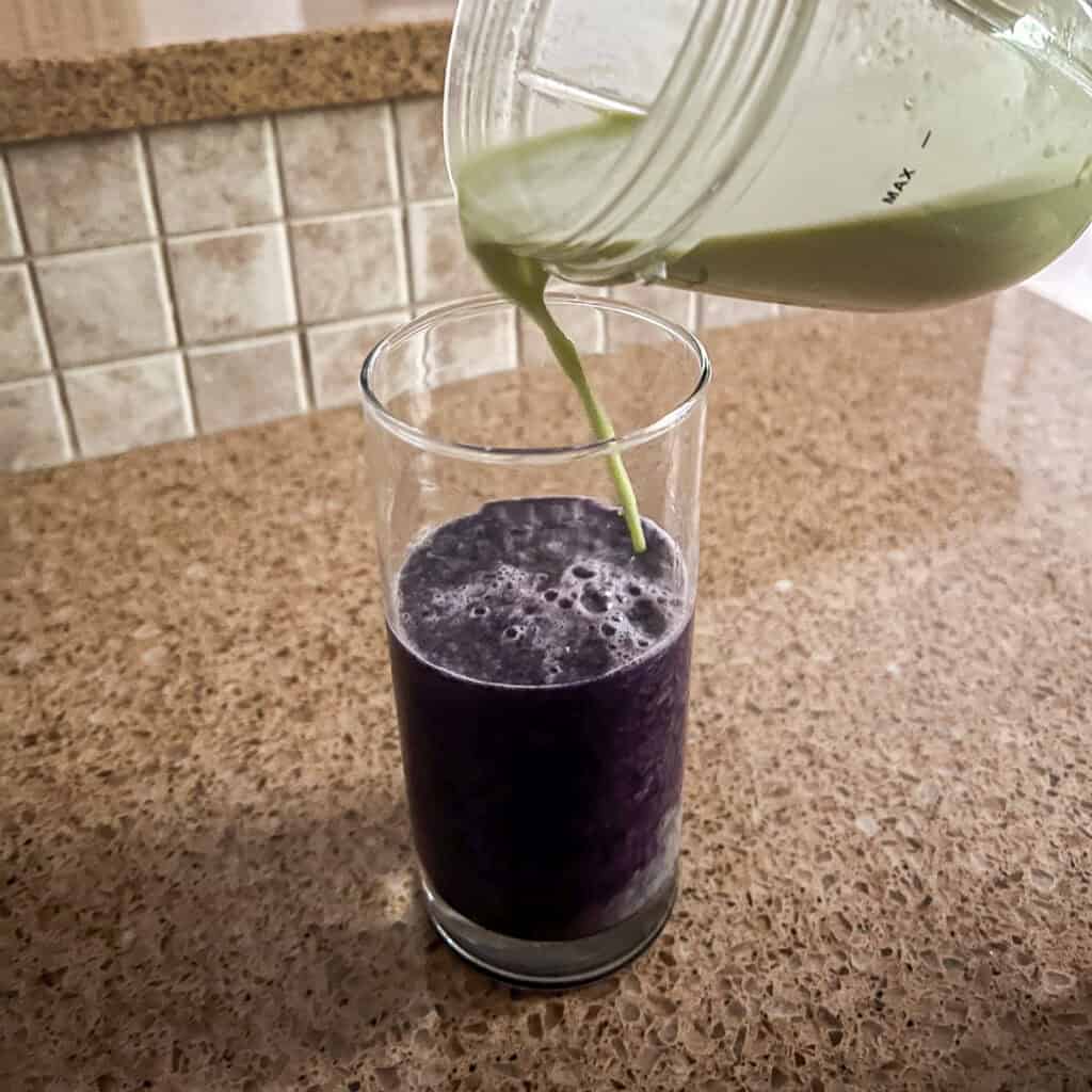A blueberry milk mixture in a tall glass with a matcha milk mixture pouring into the same glass.