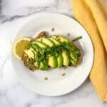 Pesto avocado toast topped with chopped spinach and roasted pine nuts on a plate with lemon wedge on the side.