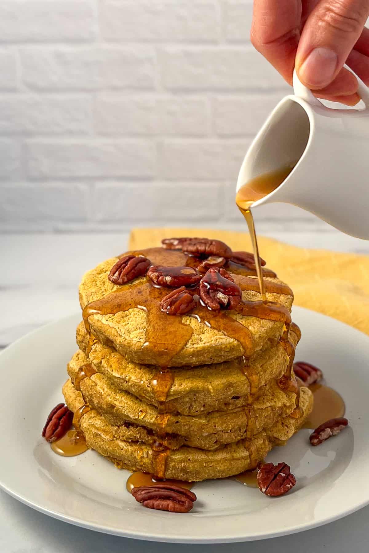 A stack of pumpkin spice pancakes topped with pecans and maple syrup being drizzled on top.