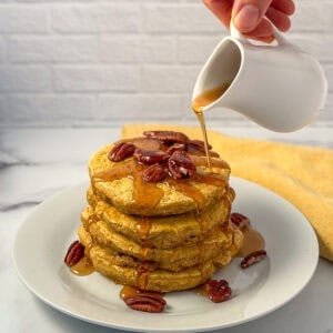 A stack of pumpkin spice pancakes topped with pecans and maple syrup being drizzled on top.