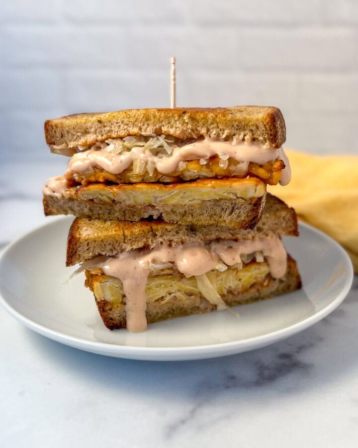 Tempeh Reuben Sandwich sliced in half on a plate with sauce dripping down.