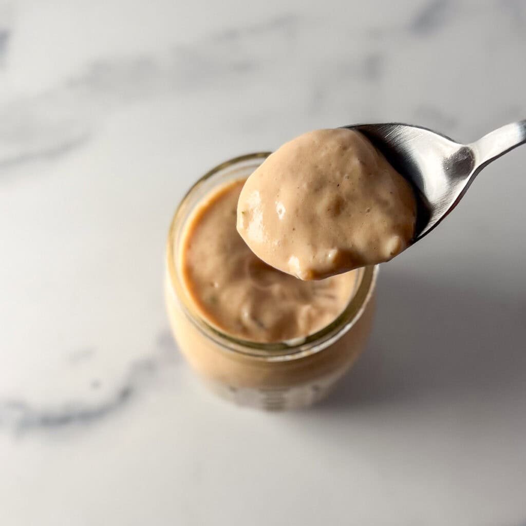 A jar of vegan thousand island dressing with a spoon lifting some out to show thick, creaminess.