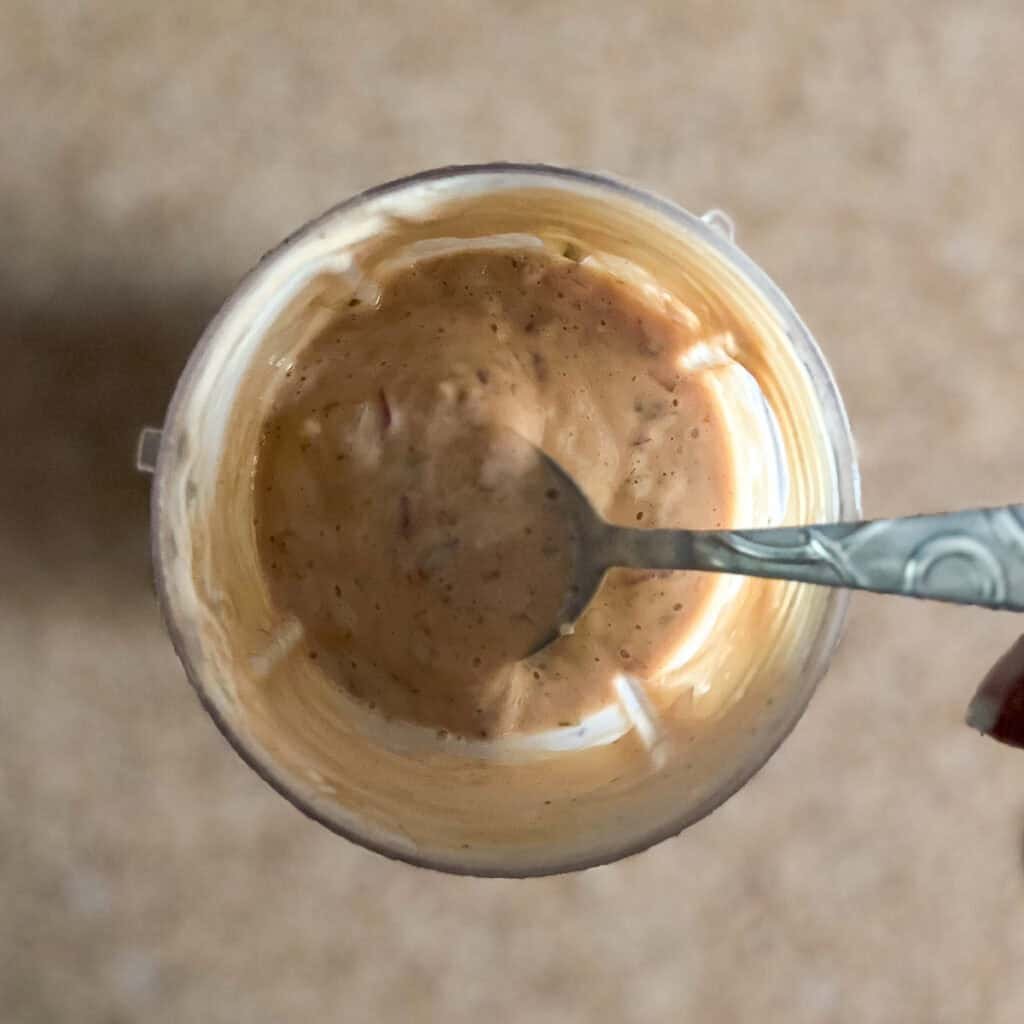 A blender with vegan thousand island dressing and a spoon stirring minced onion and relish.