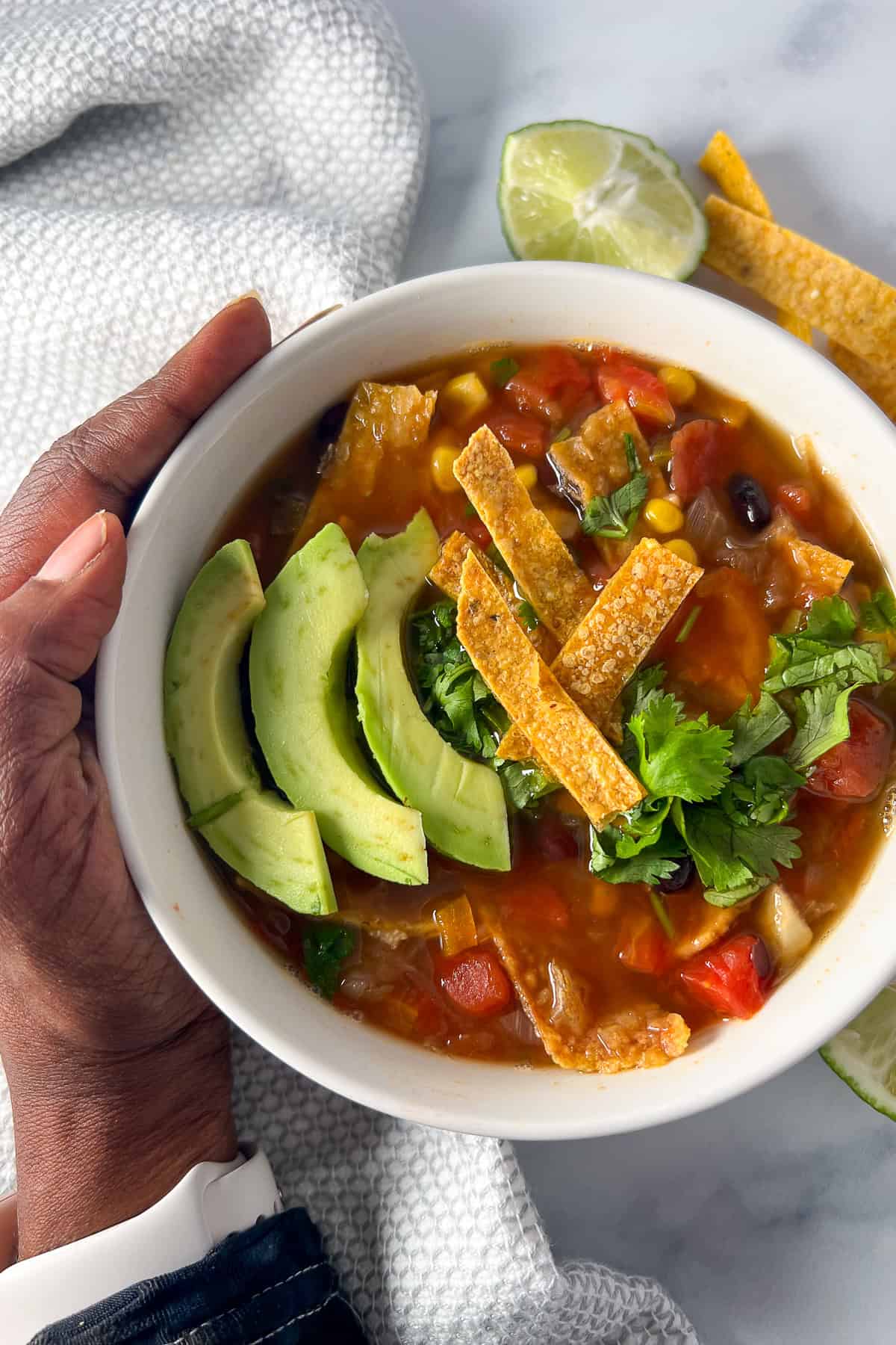 A woman's hand holding a bowl of black bean tortilla soup topped with sliced avocado; lime wedge on the side.