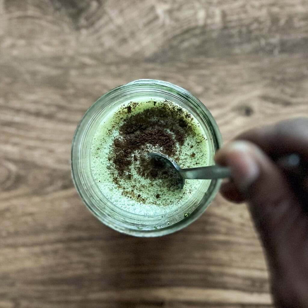 A woman's hand using a spoon to stir the vanilla bean powder and maple syrup into the matcha mixture.