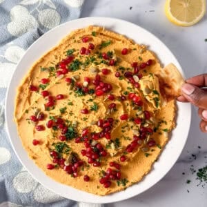 Bowl of butternut squash hummus with a woman's hand holding a pita chip dipping some out.