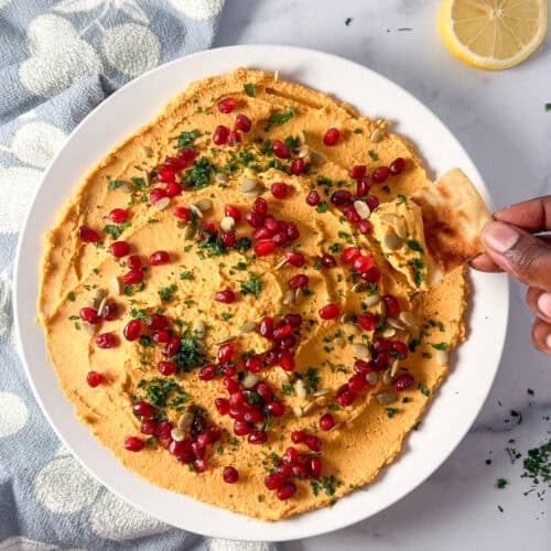 Bowl of butternut squash hummus with a woman's hand holding a pita chip dipping some out.