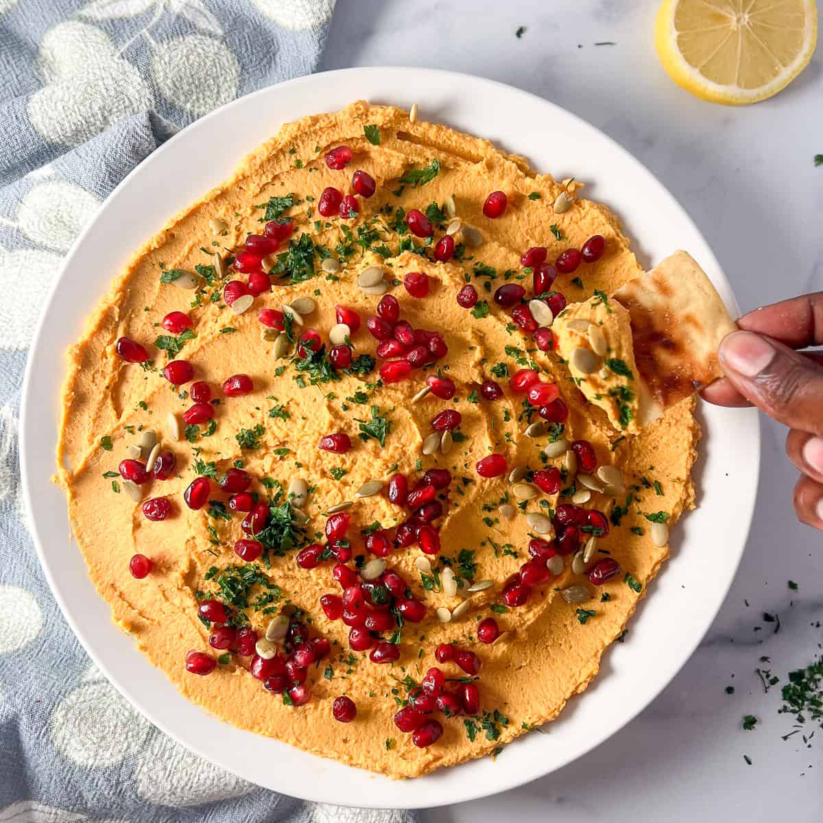 Bowl of butternut squash hummus with a woman's hand holding a pita chip dipping some out.