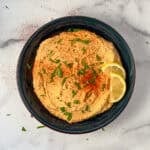 Harissa hummus in a bowl topped with fresh chopped herbs and lemon slices.