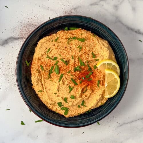 Harissa hummus in a bowl topped with fresh chopped herbs and lemon slices.