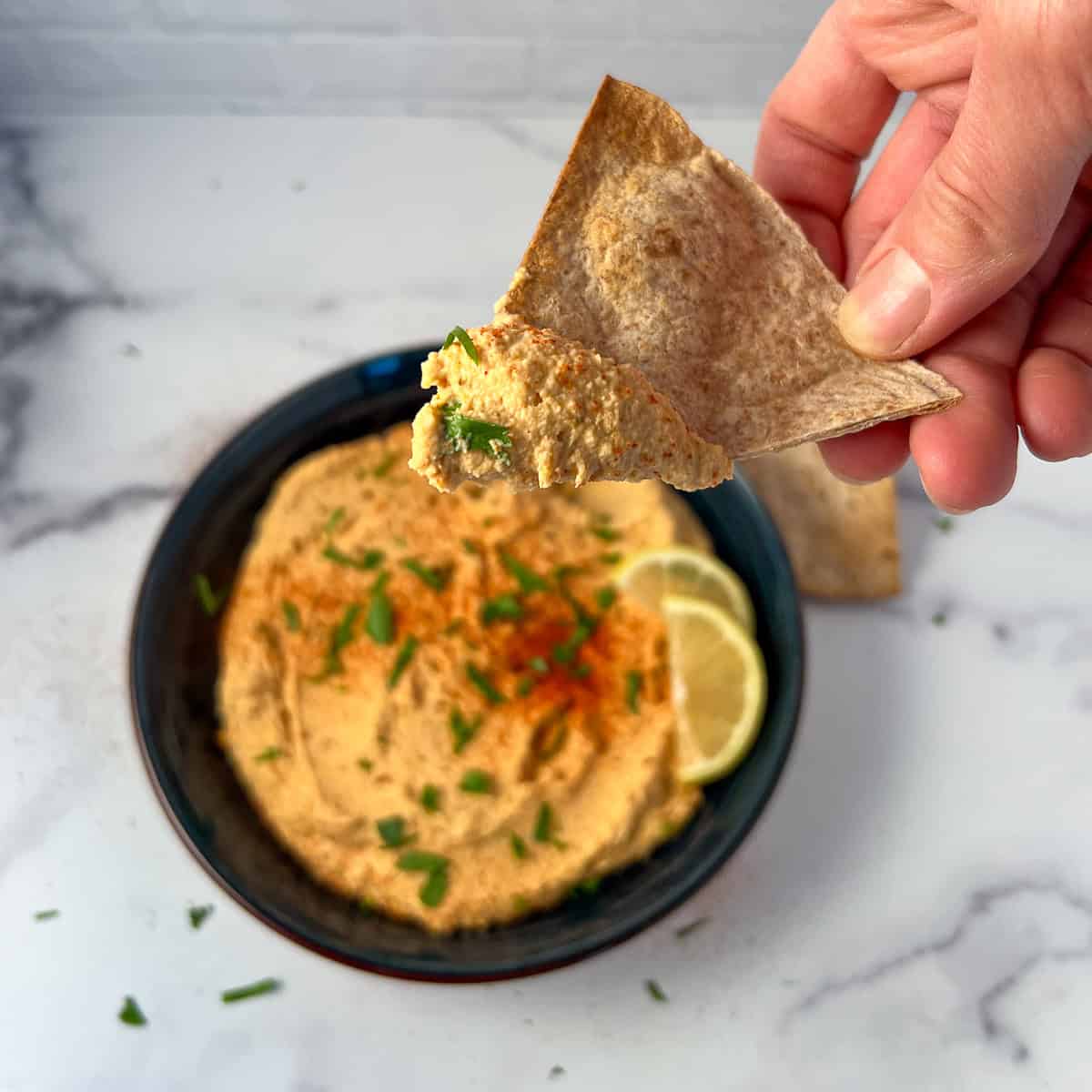 A woman's had using a pita chip to dip some harissa hummus out of the bowl.
