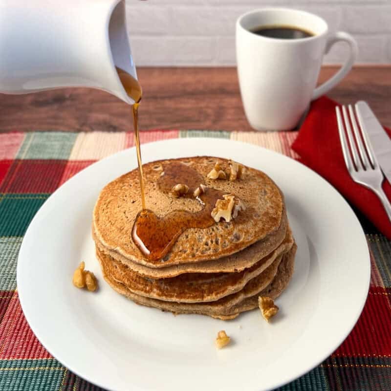 A stack of pancakes on a plate with chopped nuts and pure maple syrup being poured on top.
