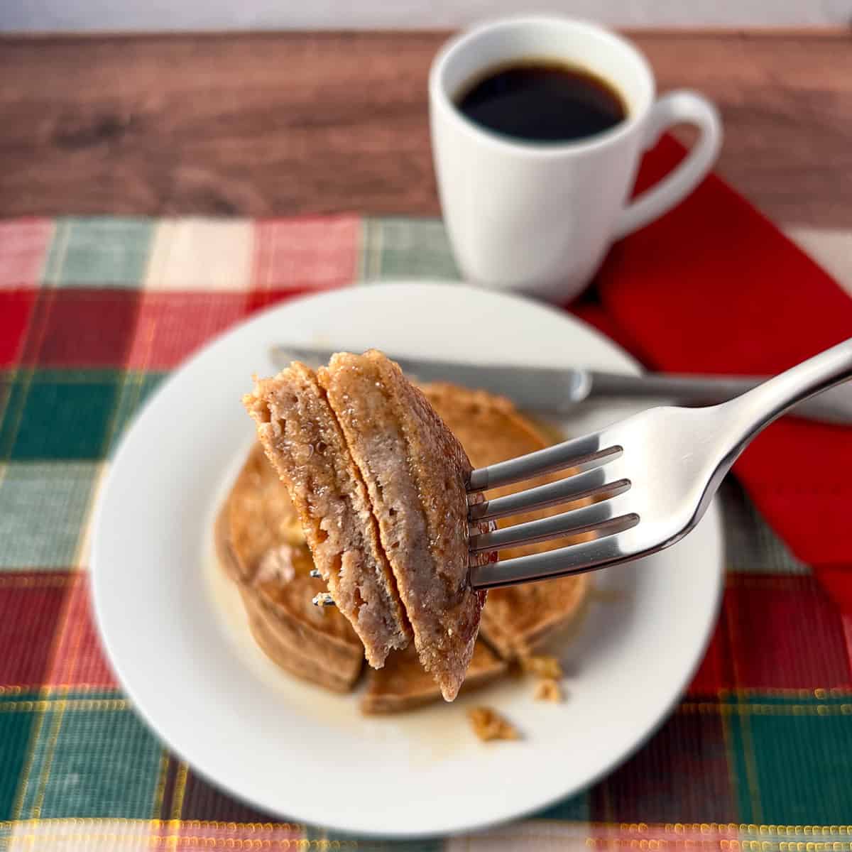 A fork with a bite of vegan eggnog pancakes with coffee in the background.