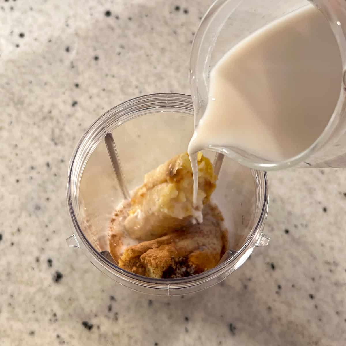Almond milk being poured into a blender cup with cooked sweet potato flesh, banana, nutmeg, vanilla and cinnamon.