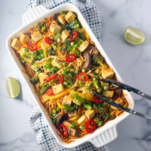 Baked Asian Noodles with veggies and tofu in a casserole dish, with lime wedges and tongs on the side.