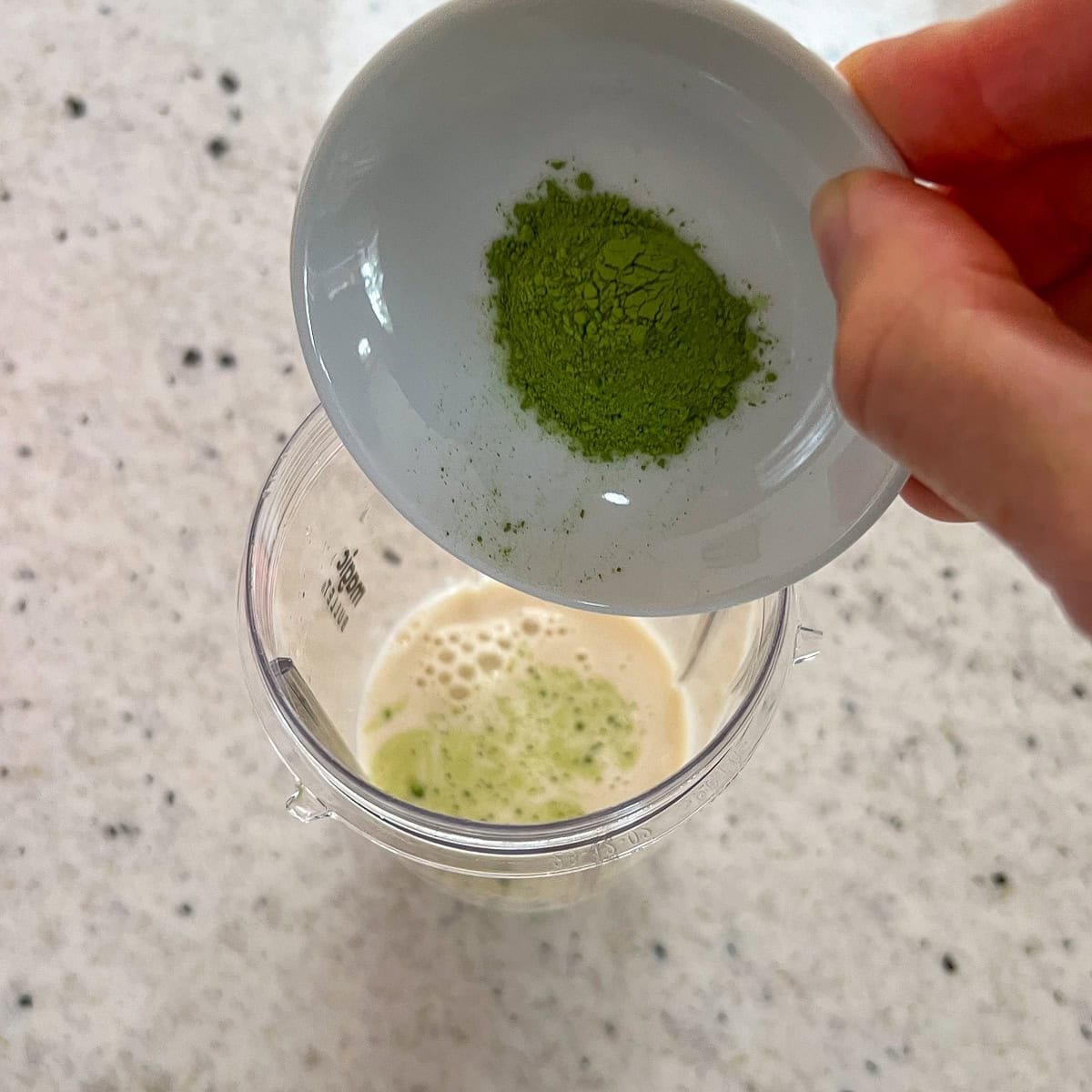 Matcha green tea powder being added to a bullet blender cup with soy milk.