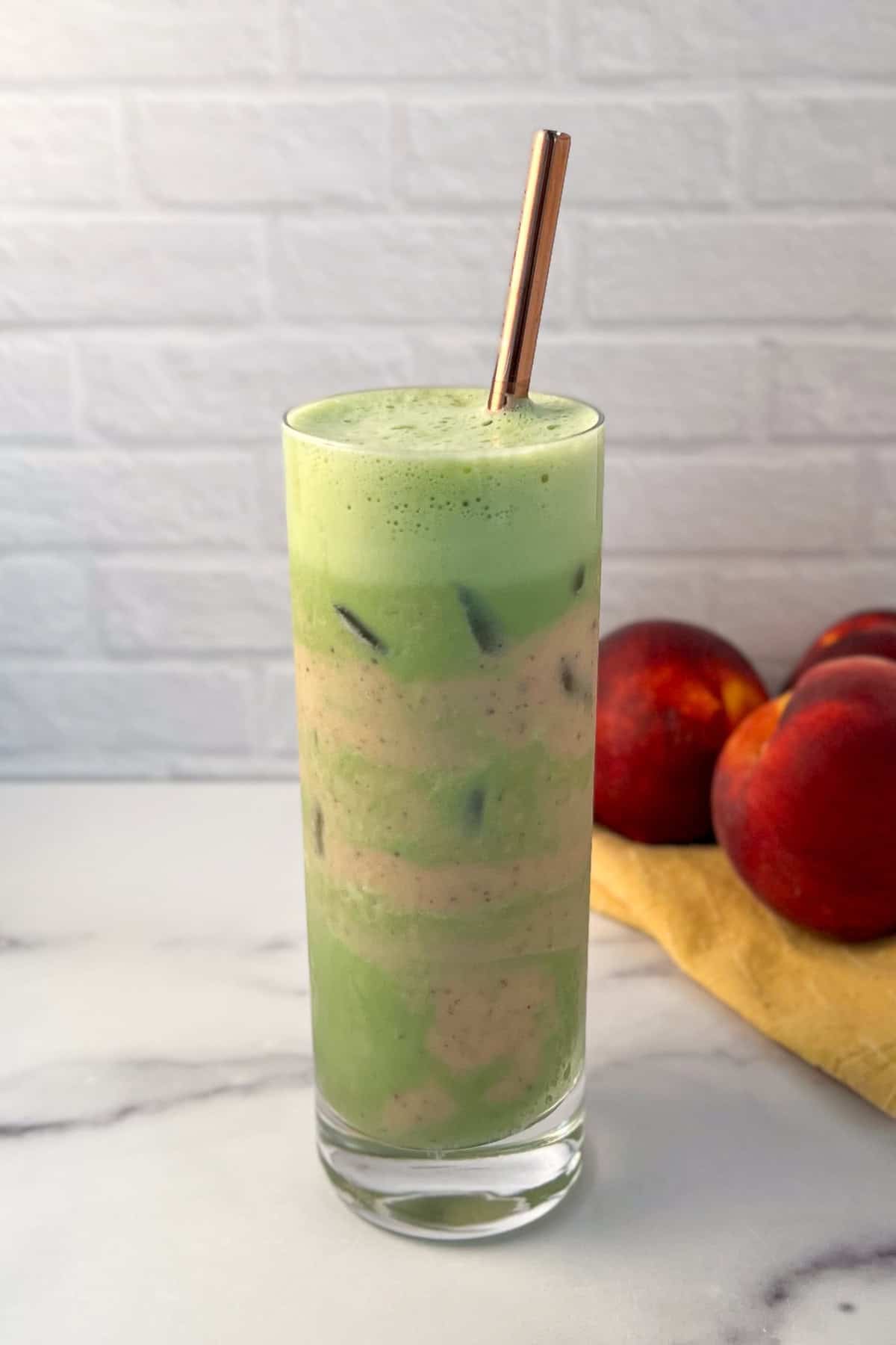 Iced peach matcha latte in a tall glass with straw; whole peaches in the background.