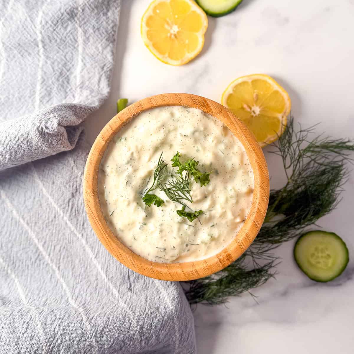 Vegan tzatziki sauce in a wooden bowl with fresh herbs and sliced lemon and cucumbers in the background.