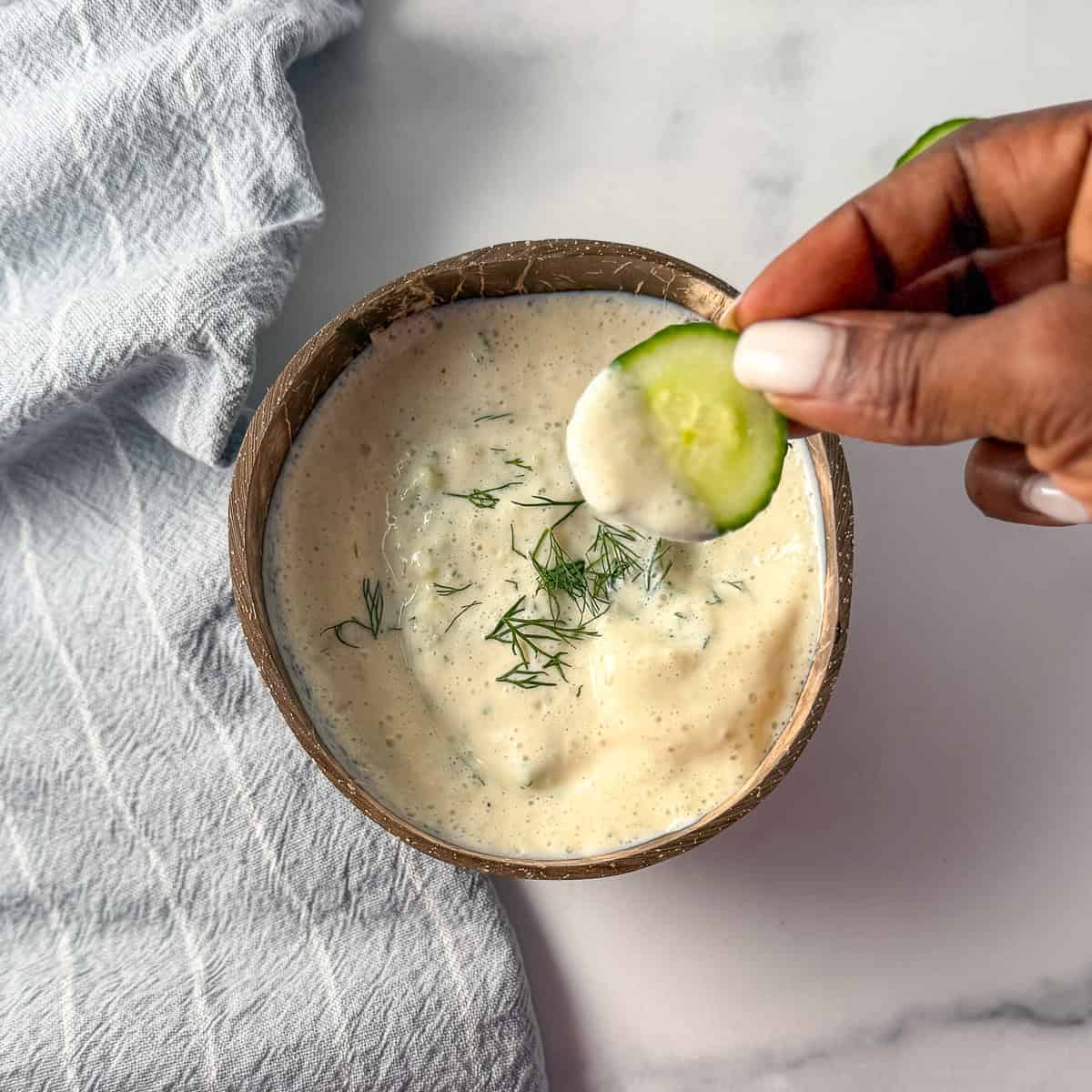 Vegan tzatziki in a bowl with a woman's hand dipping sliced cucumber in it.