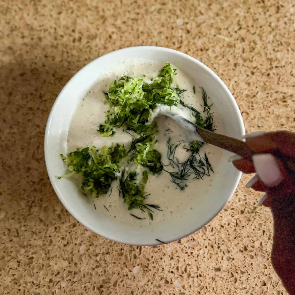 Vegan tzatziki ingredients being combined in a bowl with a spoon.