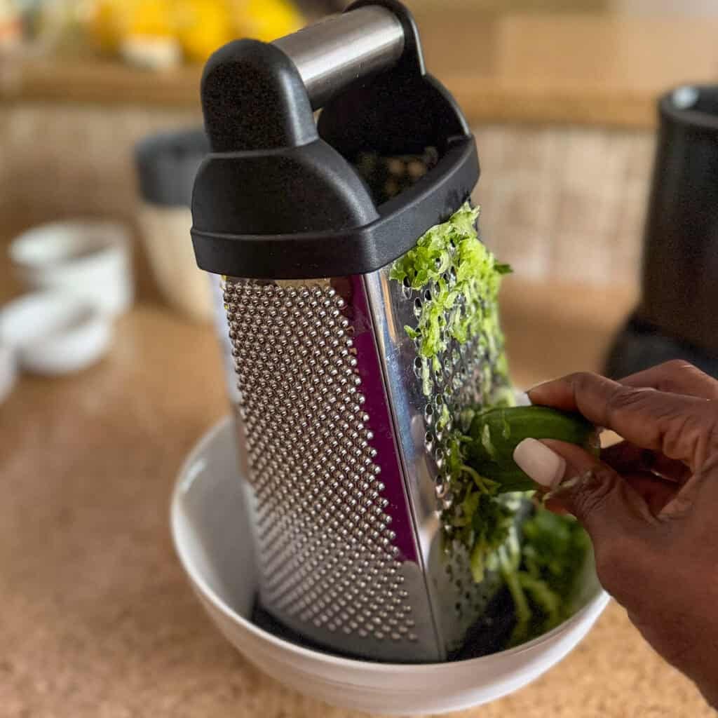 A woman's hand using a box grater to grate the english cucumber over a bowl.